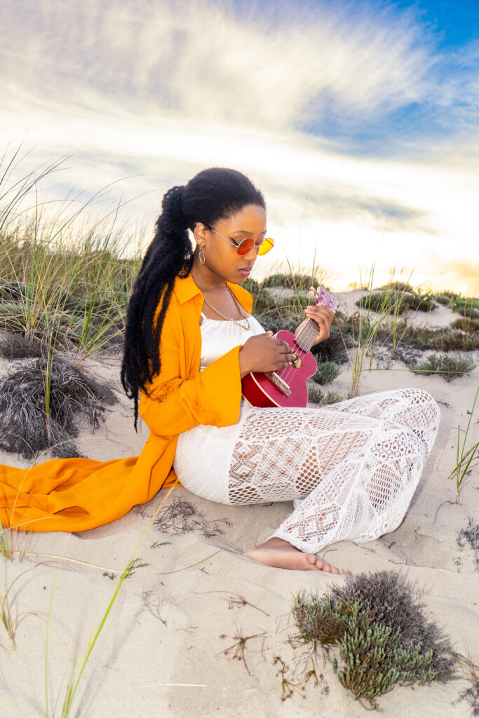 Woman on beach with orange jacket and white pants/top, playing ukulele