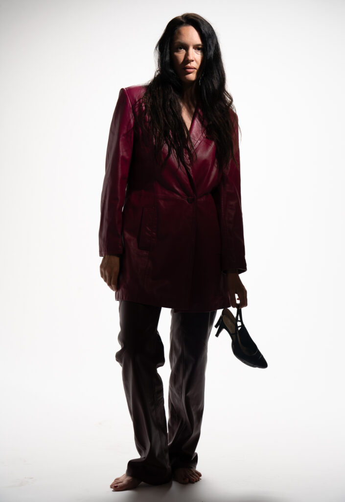 Woman in maroon leather jacket, barefoot with high heels in left hand
